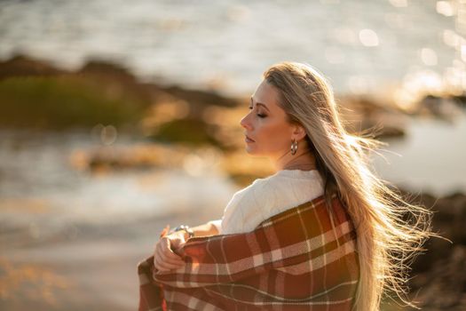 Attractive blonde Caucasian woman enjoying time on the beach at sunset, sitting in a blanket and looking to the side, with the sunset sky and sea in the background. Beach vacation