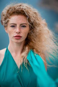 Outdoor portrait of a young beautiful natural redhead girl with freckles, long curly hair, in an emerald dress, posing against the background of the sea