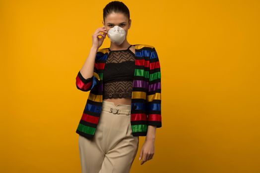 Beautiful slim girl fashion model posing in a protective respirator on a yellow background. LGBT community rainbow color jacket