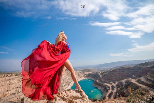 Side view of a beautiful sensual woman in a red long dress posing on a rock high above the lake in the afternoon. Against the background of the blue sky and the lake in the form of a heart.