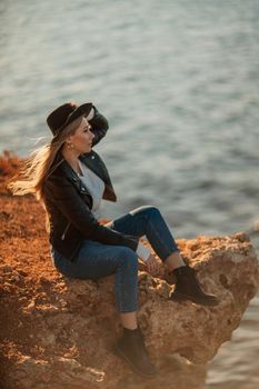 A blonde girl in a stylish black leather jacket is sitting on the seashore