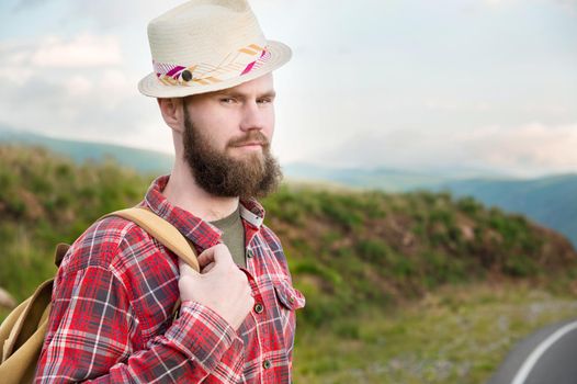 Attractive romantic bearded Caucasian man wearing a hat, plaid shirt and a backpack. Stands in the countryside in the mountains and looks at the camera. Copy space. Hitchhiker.
