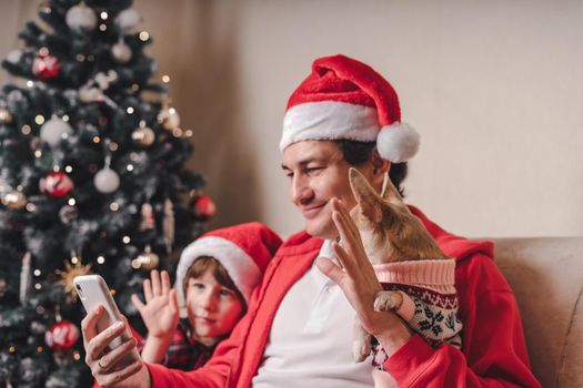 Father with child and puppy dog in Santa hats having a video call on Christmas day at smartphone, sitting on a couch in the living room with Christmas tree at home.