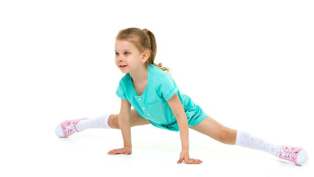 Adorable active little girl doing split on floor. Adorable girl dressed in cotton jumpsuit, white knee socks and sneakers sitting on twine on isolated white background in studio