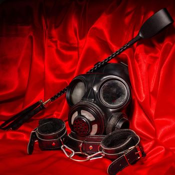 Close up bdsm outfit. Bondage, kinky adult sex games, kink and BDSM lifestyle concept with gas mask, whip, collar, leather handcuffs on red silk with copy space.