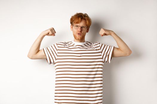 Image of confident and strong redhead man flexing biceps, showing muscles after gym, standing over white background.