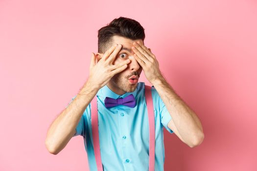 Intrigued funny guy in bow-tie, peeking through fingers at something interesting, checking out something cool, standing on pink background.