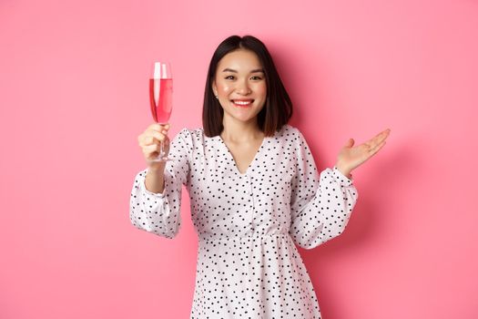 Beautiful asian woman raising glass of champagne and looking happy at camera, congratulating you on party, celebrating, standing over pink background.