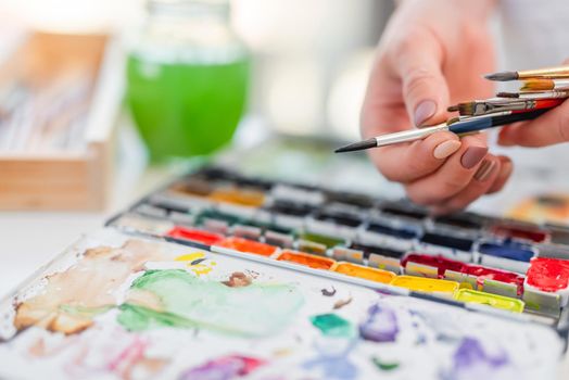 Artist woman holding different painbrushes in her hands and choosing one with colourful watercolor on background