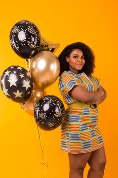 Portrait of smiling young African-American adult woman looking sweet on yellow background holding balloons.