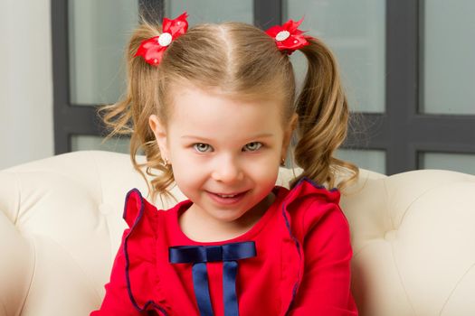 Cute blonde little girl sitting on comfy armchair. Charming lovely kid with cute ponytails hairstyle dressed red dress.Close up