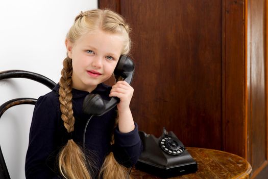 Blonde girl talking by old black phone. Lovely girl with braids sitting on chair in vintage room interior with old wooden furniture. Cute six years old kid speaking on retro telephone