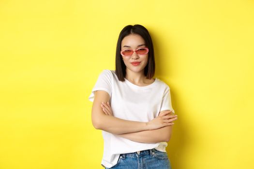 Fashion and lifestyle concept. Sassy and confident asian woman in trendy sunglasses looking self-assured at camera, standing over yellow background.