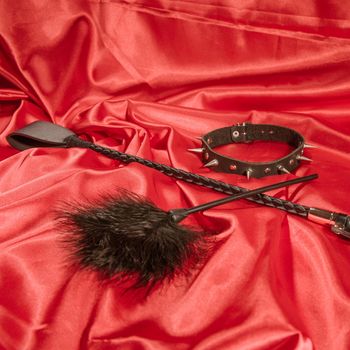 Bondage, kinky adult sex games, kink and BDSM lifestyle concept with a whip, feather stick, collar on red silk with copy space