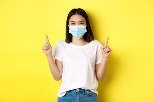 Covid-19, pandemic and social distancing concept. Young asian woman in white t-shirt and medical mask from coronavirus, pointing fingers up, showing special offer.