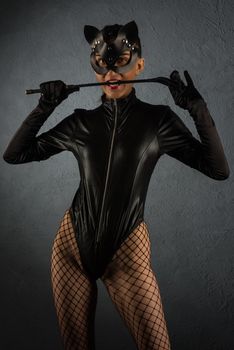 Adult sex games. Beautiful dominant brunette vamp mistress girl in latex body, gloves and bdsm black leather fetish cat mask posing with riding crop. - image