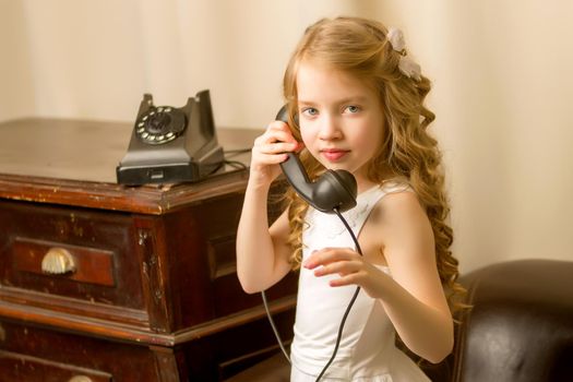 A beautiful little girl in a room in the fifties of the last century, talking on an old phone with a big black tube. Retro style, black and white photo in the studio. Concept of nostalgia, vintage.
