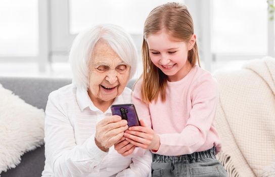 Grandmother with grandaughter sitting on the sofa at home and looking at smartphone with smile