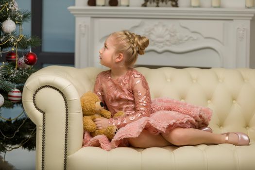 Cute little girl on Christmas night, sitting on a sofa near the New Year trees with a teddy bear in her hands. Family holidays concept.