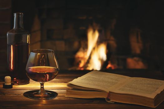 Glass of cognac, a bottle and an open old book on the table near the burning fireplace. Rest and relaxation concept.