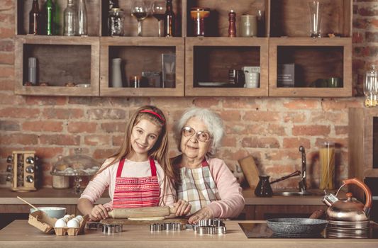 Eldery granny with her lovely granddaughter roll out the dough using the rolling pin and looking at camera