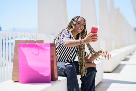 Young black woman taking a selfie with her smartphone sitting on a bench in the street.