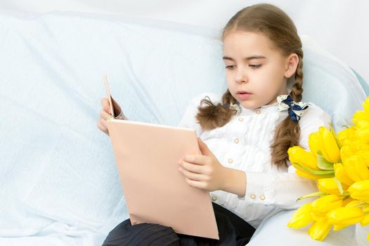 A beautiful little girl with long blond, flowing hair sits on the couch next to a large bouquet of yellow flowers, tulips.She is reading a book.