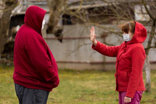 An elderly woman in a protective medical mask shows a gesture of her hand about keeping the social distance of a senior man, standing in front of an outdoor street.