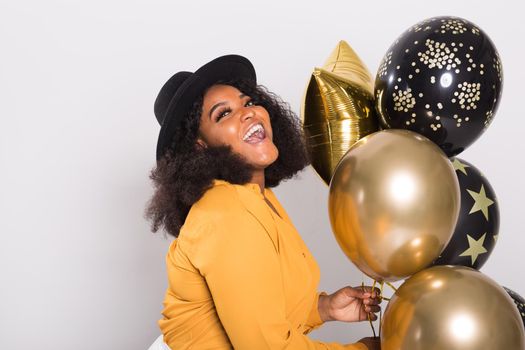 Portrait of smiling young African-American adult woman looking sweet on yellow background holding balloons.