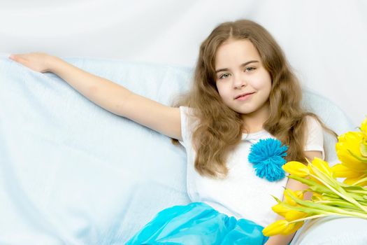 A beautiful little girl with long blond, flowing hair sits on the couch next to a large bouquet of yellow flowers, tulips.