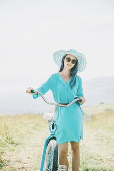 Beautiful fashionable young woman wearing in blue dress and hat walking with bicycle on summer meadow, looking at camera.