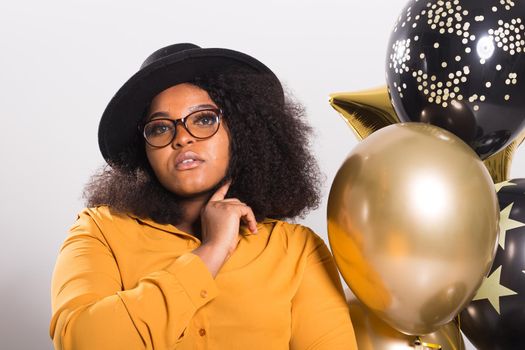 Portrait of smiling young African-American adult woman looking sweet on yellow background holding balloons.