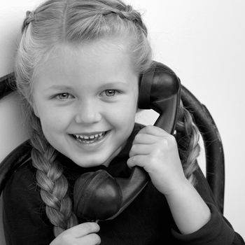 Smiling girl talking by old phone. Black and white shot of lovely kid sitting on chair in vintage room interior. Cute six years old kid speaking on vintage telephone