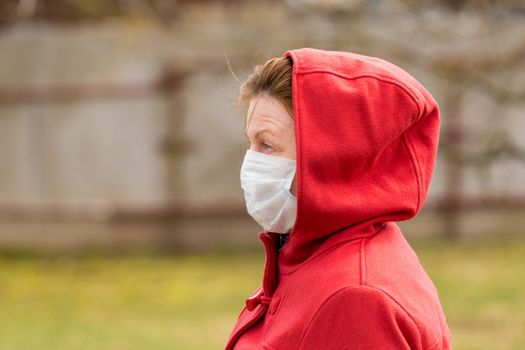 An elderly woman with brown hair and blue eyes in a red coat and hood in a protective safe medical mask, close-up portrait.