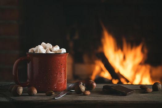 Cocoa with marshmallows and chocolate in a red mug on a wooden table near a burning fireplace.