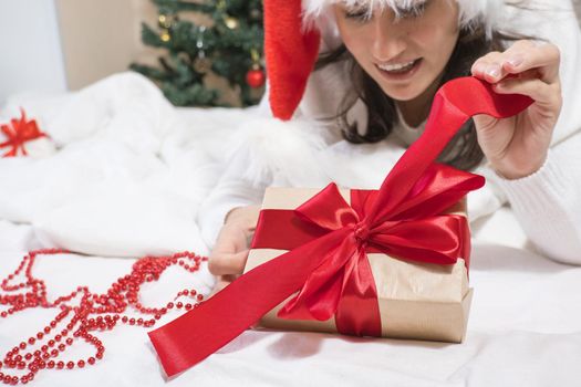 Happy young brunette woman in santa claus hat opens a gift box for Christmas lying on a white bed. Christmas gifts