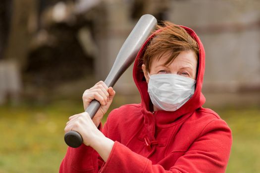 Angry aggressive elderly woman in protective safe medical mask swings baseball bat in the background of outdoor street, portrait, close up.