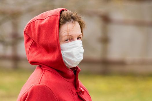 An elderly woman with brown hair and blue eyes in a red coat and hood in a protective safe medical mask, close-up portrait.