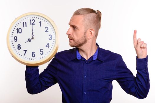 Young handsome man in blue shirt looks at big clock standing pointing upwards on white background.