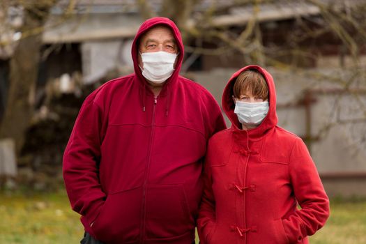 An elderly man and woman Caucasian or European hooded look stand next to each other in an open-air street, in protective safe medical masks against coronavirus.
