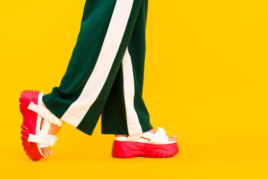 Women's legs in sport sandals with red soles and green pants with stripes on a yellow background.