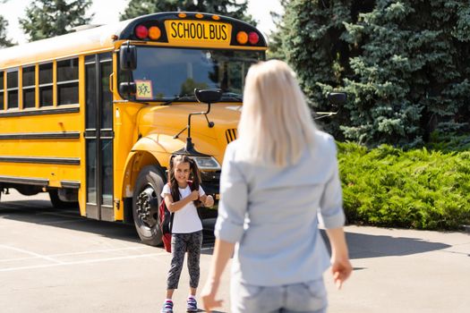 Mother escorts the schoolgirl with ponytails and a backpack to school.