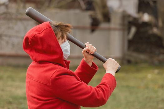 Angry aggressive elderly woman in protective safe medical mask swings baseball bat in the background of outdoor street, portrait, close up.