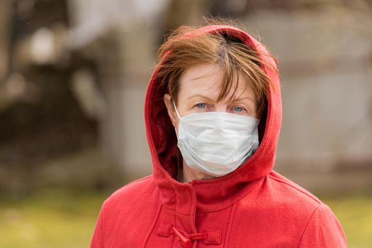 An elderly woman with brown hair and blue eyes in a red coat and hood in a protective safe medical mask, close-up portrait.