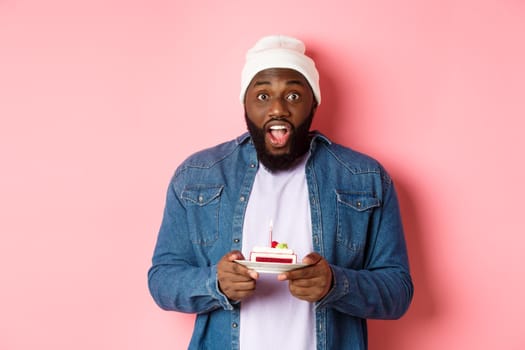 Excited Black man celebrating birthday, staring at camera amazed while making wish on bday cake with candle, standing over pink background.