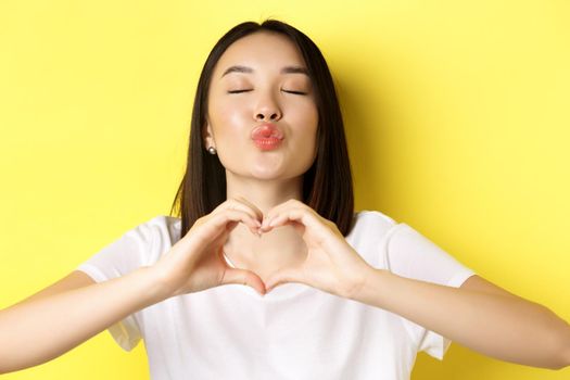 Valentines day and women concept. Close up of pretty asian girl in white t-shirt, smiling and showing heart, I love you gesture, standing over yellow background.