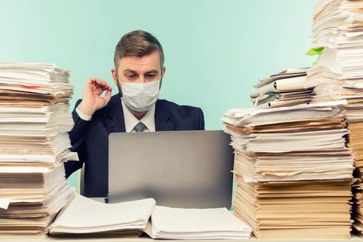 A male accountant or company manager works in an office in a pandemic in view of the accumulated paper work. A protective medical mask is on the face. On the desktop are large stacks of documents. - image