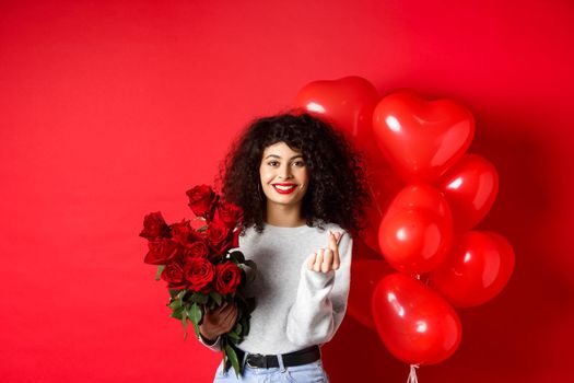 Holidays and celebration. Beautiful girlfriend receive flowers on anniversary, showing finger heart and standing near party balloons, red background.