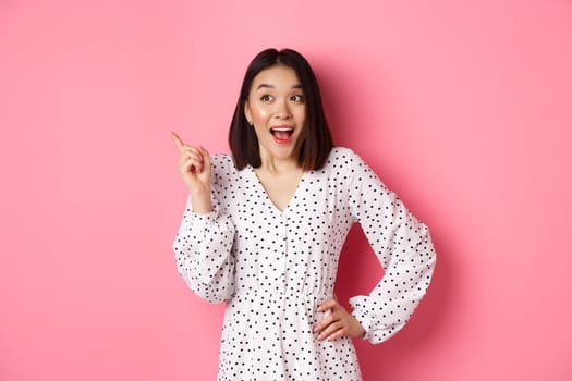 Excited asian woman in stylish dress, gasping amused, pointing finger upper left corner, showing interesting promo offer, standing over pink background.