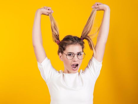 A young girl in glasses holds herself by the tail and screams isolated on a yellow background